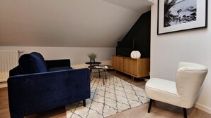 A living room with a blue sofa, a white armchair, a small table with a plant, and a framed picture on the wall. - The KAT106 - 1 bedroom (Liège)
