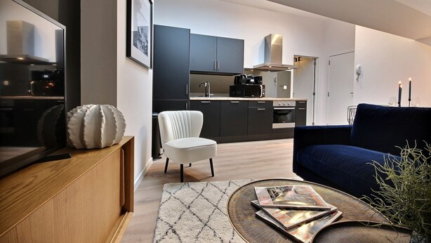 A modern living room with a dark blue sofa, a white armchair, and a coffee table with magazines. A kitchen with dark cabinets and a microwave is visible in the background. - The KAT106 - 1 bedroom (Liège)
