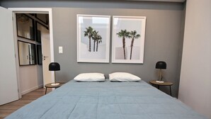 A bedroom with a bed, two bedside tables with lamps, a door leading to a bathroom, and two framed pictures of palm trees on the wall. - The KAT106 - 1 bedroom (Liège)