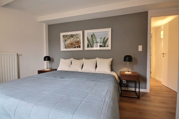 Een slaapkamer met een groot bed, twee nachtkastjes met lampen, een radiator en twee ingelijste foto’s aan de muur.