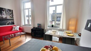 A modern living room with a red sofa, a glass coffee table, a dining area with a table set for two, and a large window with a city view. - Jardin Botanique - 1 bedroom (Liège)