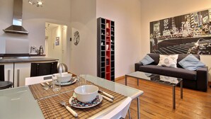 A modern living room with a glass dining table, a sofa with cushions, and a cityscape painting on the wall. - Jardin Botanique - 1 bedroom (Liège)