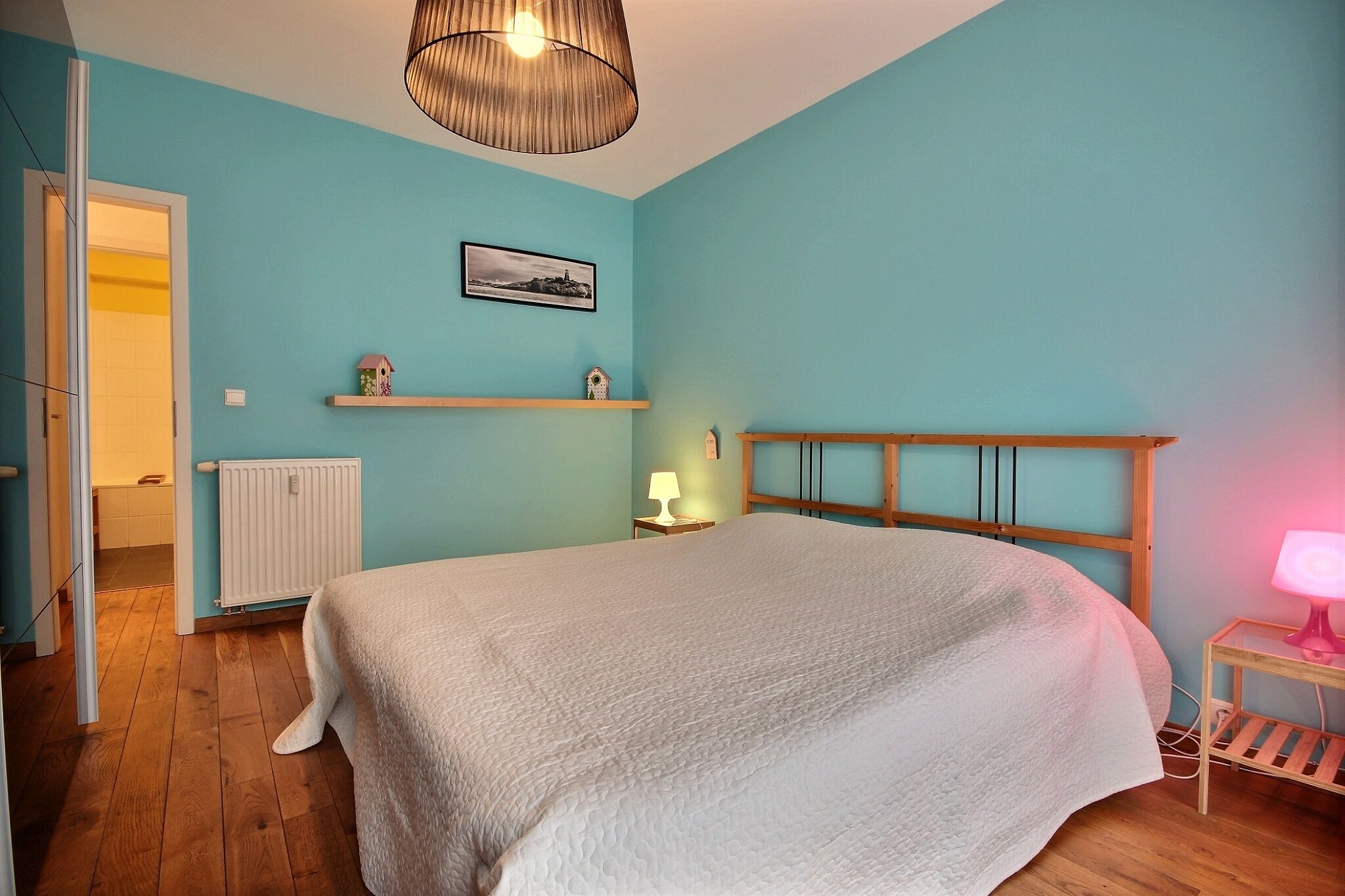 A bedroom with a large bed, a wooden headboard, a radiator, a shelf with decorative items, and a hanging light fixture.