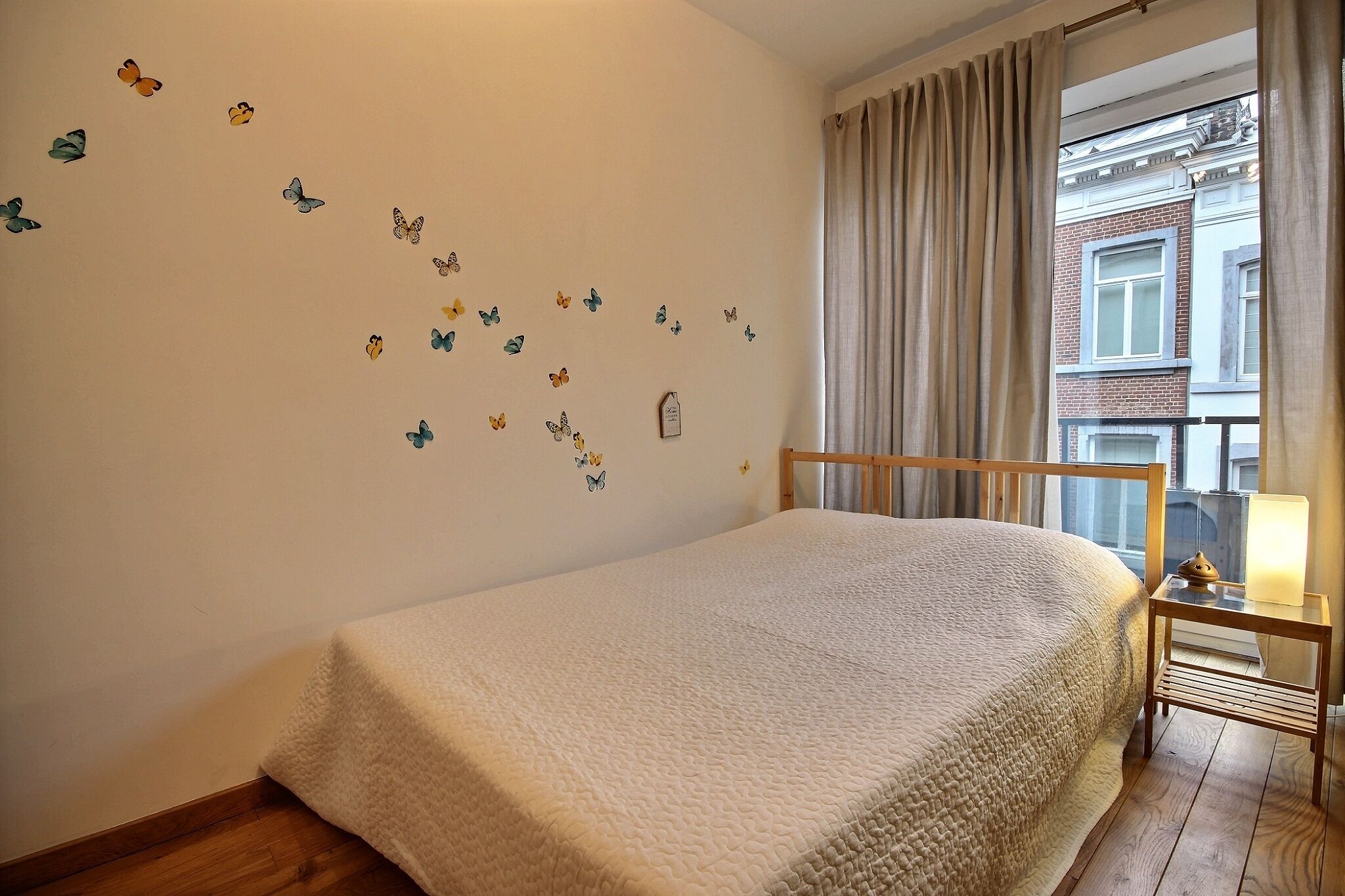 A bedroom with a bed, a nightstand with a lamp, a window with curtains, and butterfly decals on the wall.