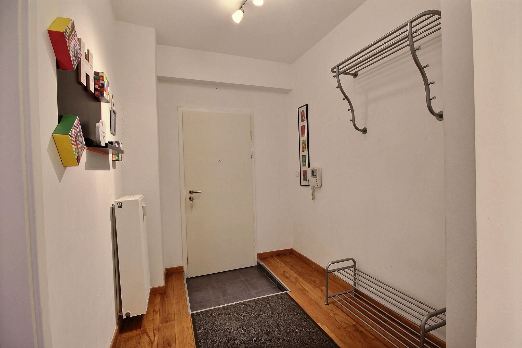 Een gang met een radiator, een deur, een plank met kleurrijke objecten en een metalen rek.