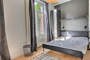 Ein Schlafzimmer mit Bett, Nachttisch, Lampe, Heizkörper, einem Fenster mit Vorhängen und Blick ins Freie.