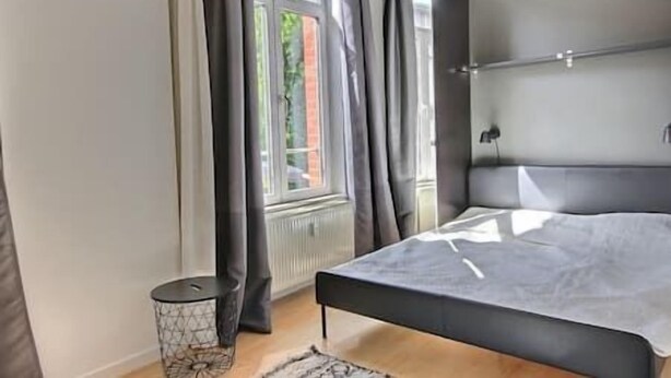 A bedroom with a bed, a radiator, a window with curtains, and a small round table.