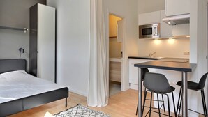 A compact bedroom with a bed, a bedside table, and a hanging light fixture. Adjacent is a kitchenette with a microwave, a sink, and a small dining area with two chairs.