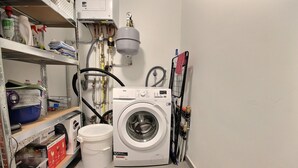 Een kleine wasruimte met een voorlader wasmachine, een boiler en diverse schoonmaakmiddelen op planken.