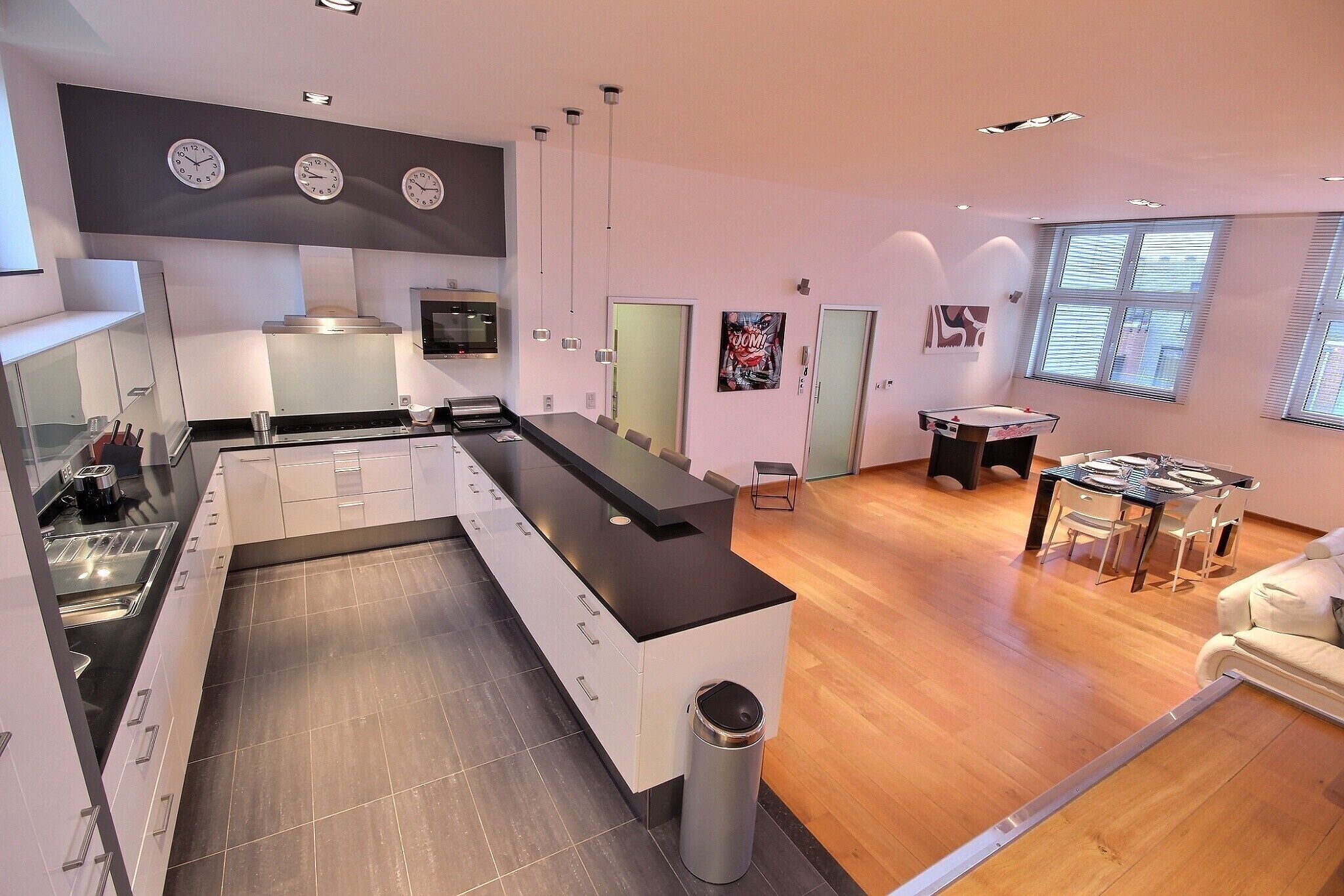 A modern kitchen with a black countertop, white cabinets, and a stainless steel sink. There is a dining area with a table and chairs, and a trash can is visible near the kitchen.