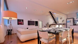Una sala de estar moderna con un sofá blanco, una mesa de comedor con sillas, una lámpara de pie y una escalera.