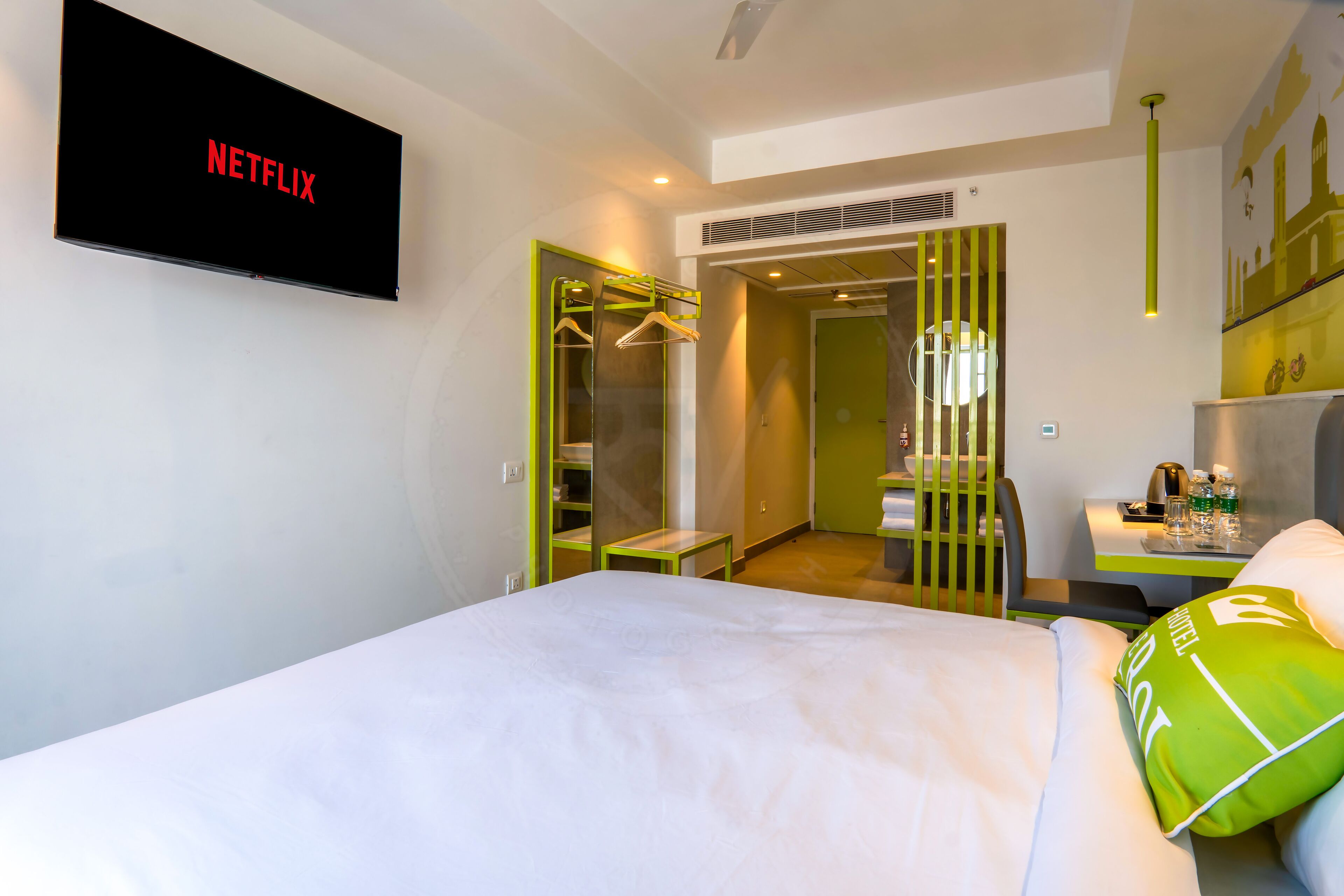 A hotel room with a bed, a TV displaying 'NETFLIX', a wardrobe, a desk with a chair, and a bathroom visible through an open door.
