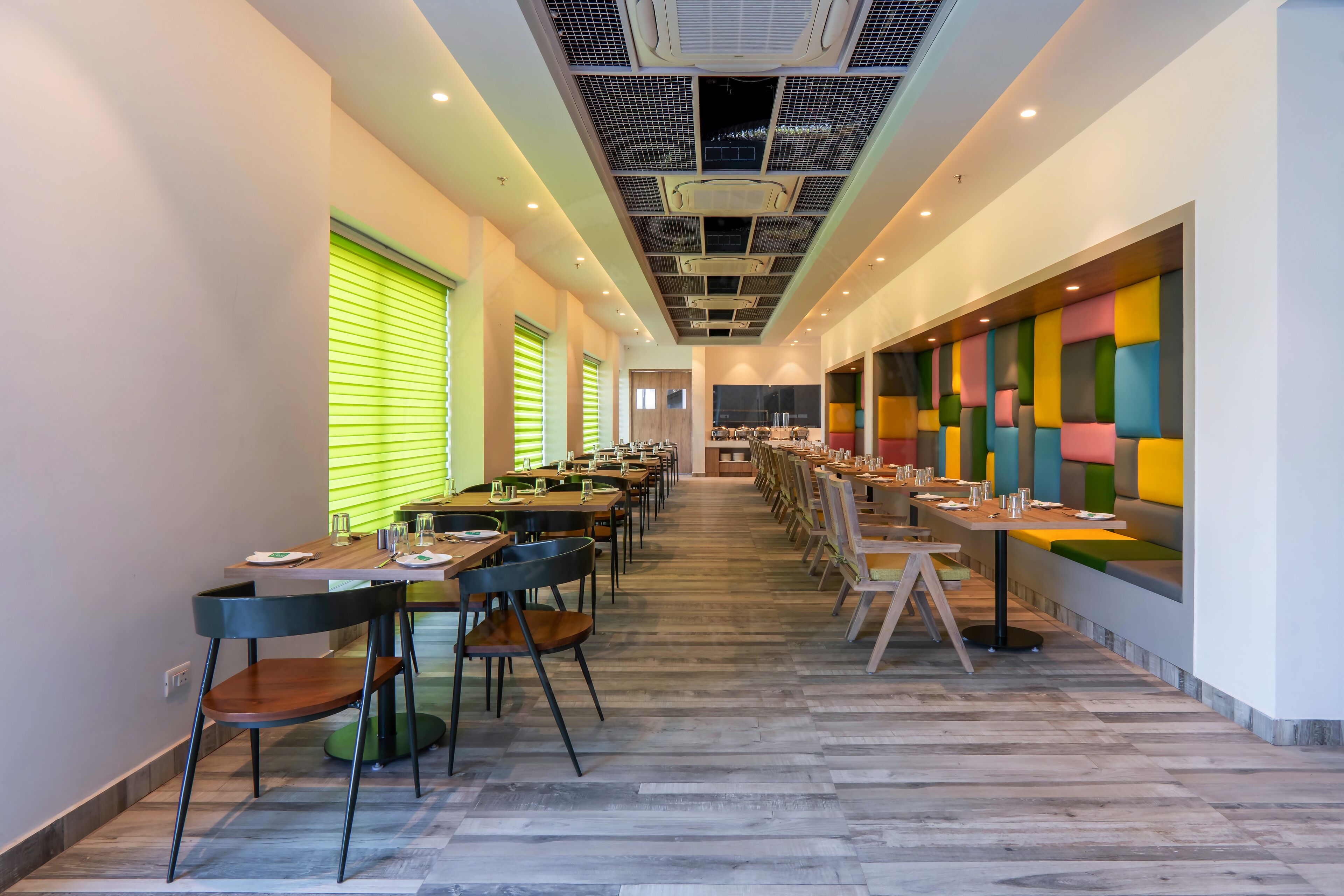 A modern dining area with long tables and chairs, a ceiling with air conditioning units, and a colorful wall design.