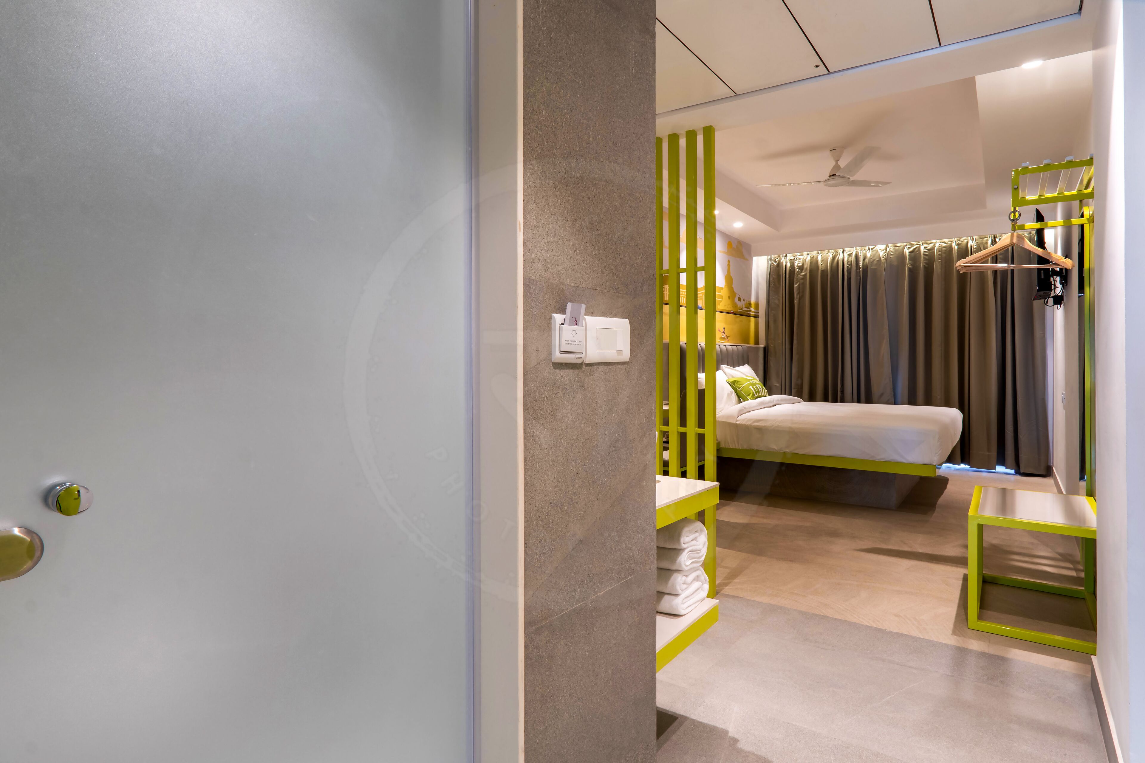 A modern hotel room with a bed, a bedside table, a hanging rack, and a wall-mounted light switch.