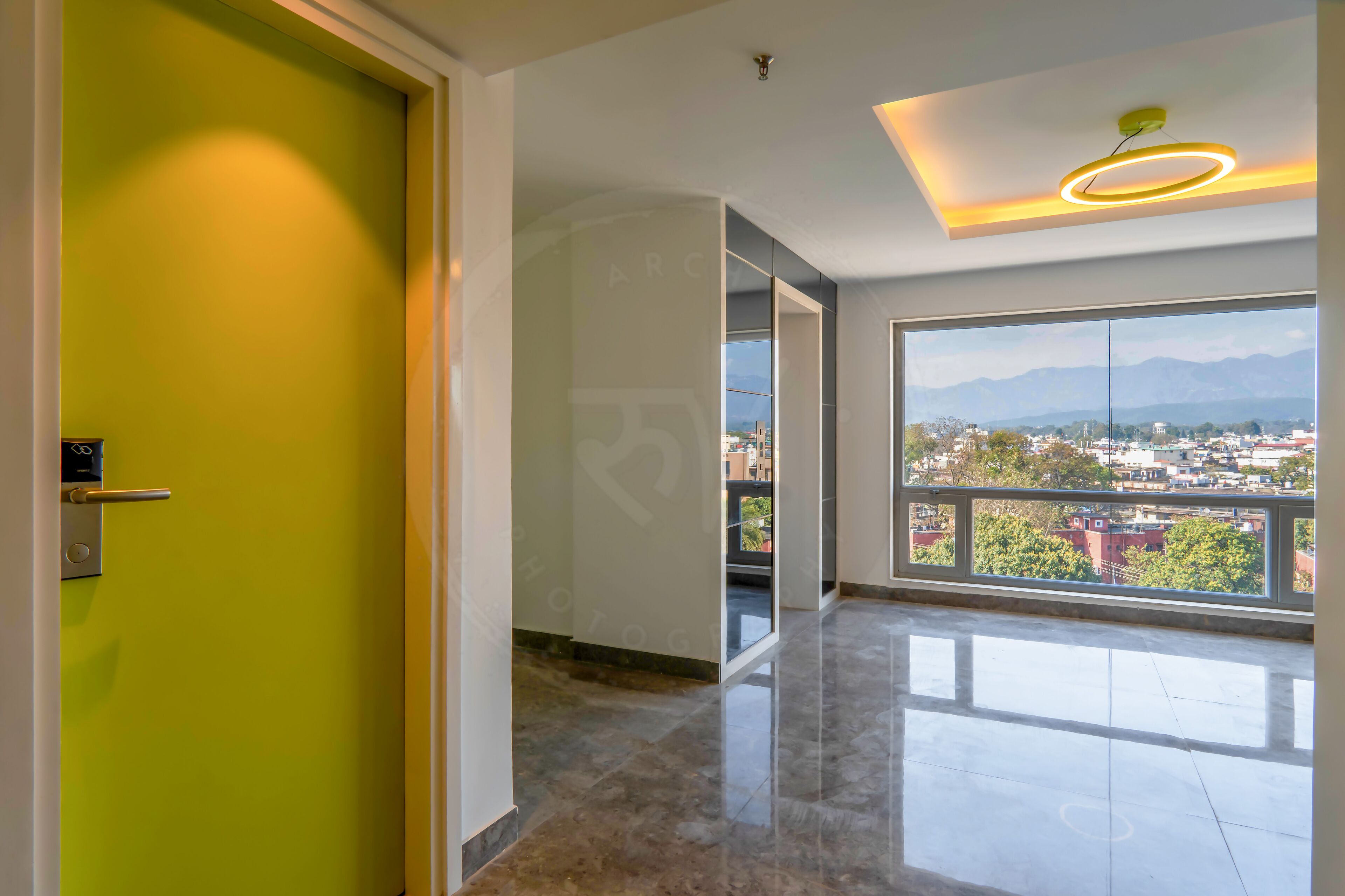 A modern hotel room with a large window offering a city view, a bright green door, and a contemporary light fixture.