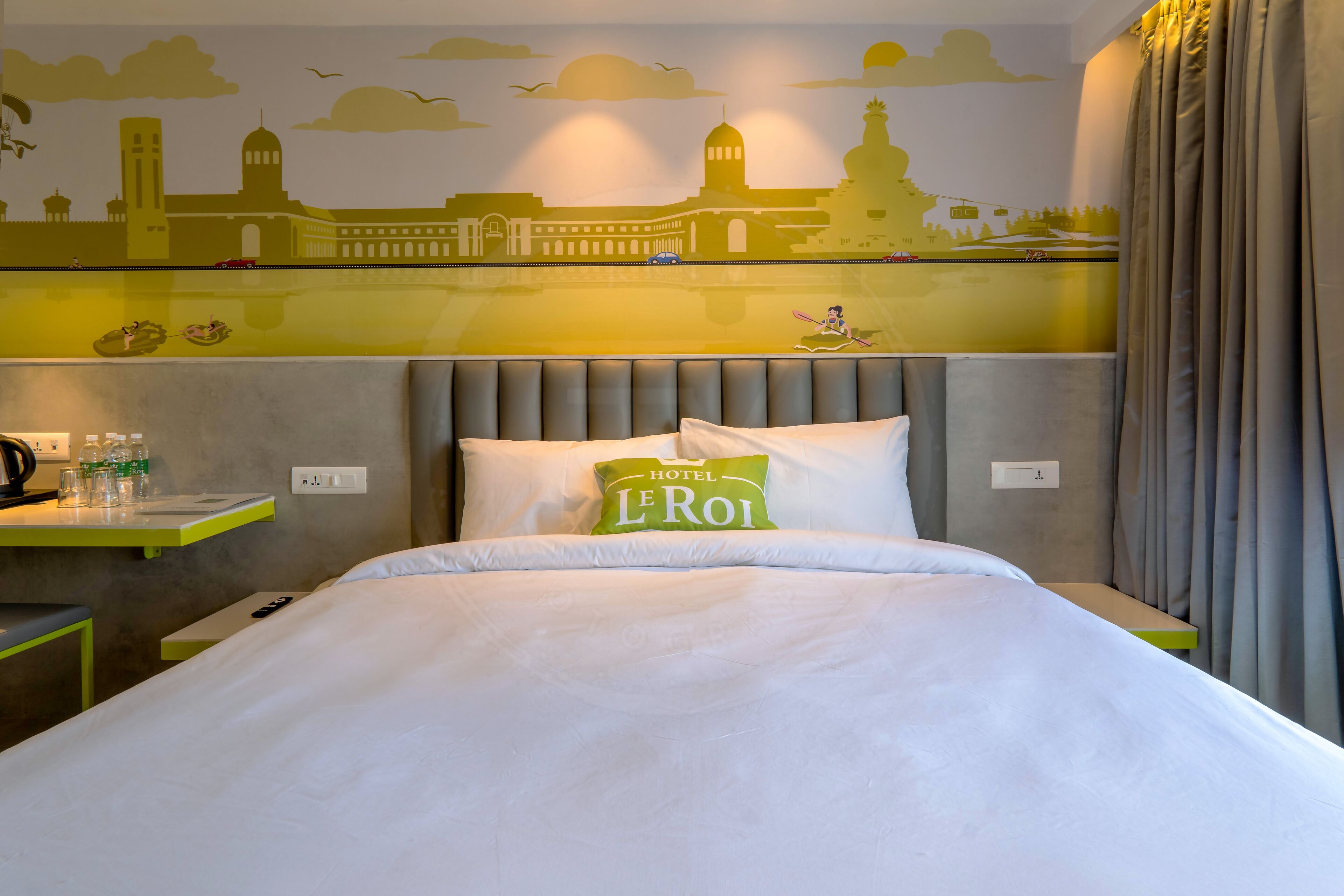 Une chambre d’hôtel avec un lit, un oreiller portant le nom de l’hôtel, une table de chevet avec une lampe et une fresque murale.
