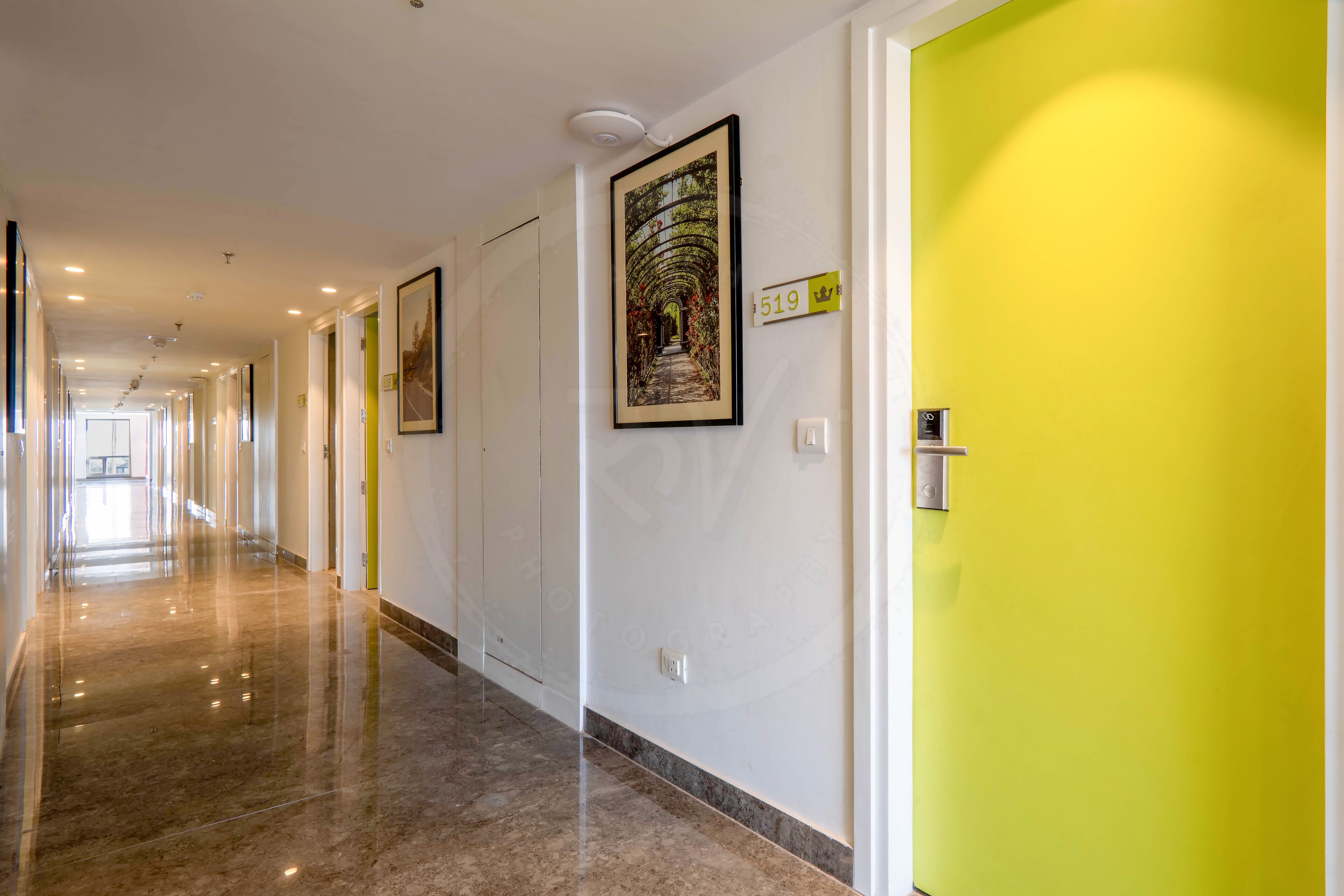 A hotel corridor with a bright yellow door, a framed picture on the wall, and a number sign.
