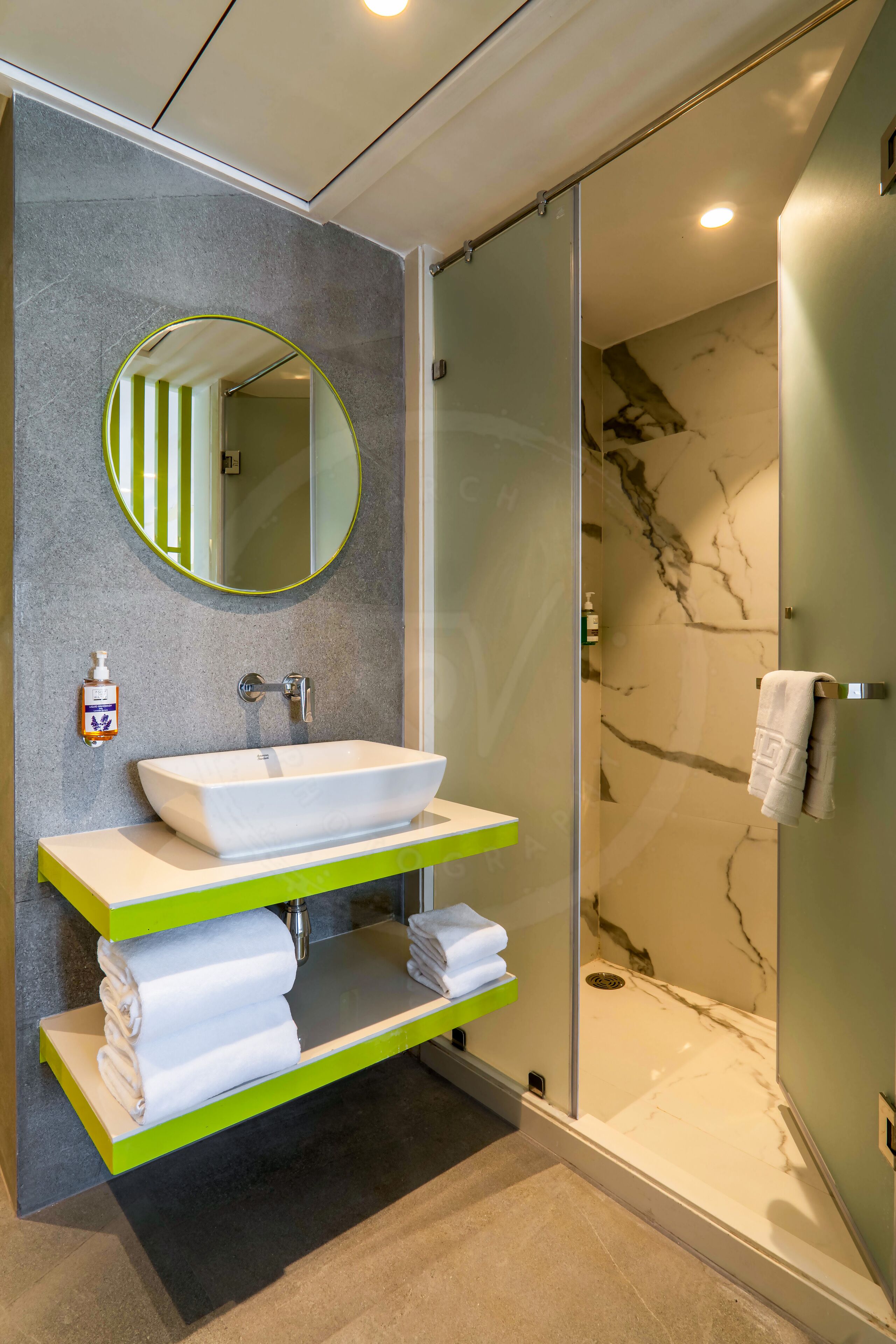 A modern bathroom with a white vessel sink, green shelves, a round mirror, and a glass-enclosed shower.