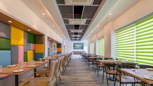 Un restaurante moderno con largas mesas de madera y asientos acolchados de colores, ventanas amplias con persianas verdes e iluminación empotrada en el techo.