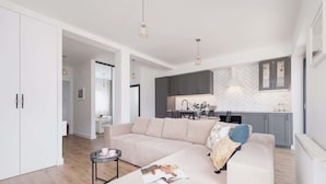 Una sala moderna con un sofá seccional beige, una mesita auxiliar y una revista sobre el sofá. Se observa una zona de cocina con armarios grises y un salpicadero tipo espiga.