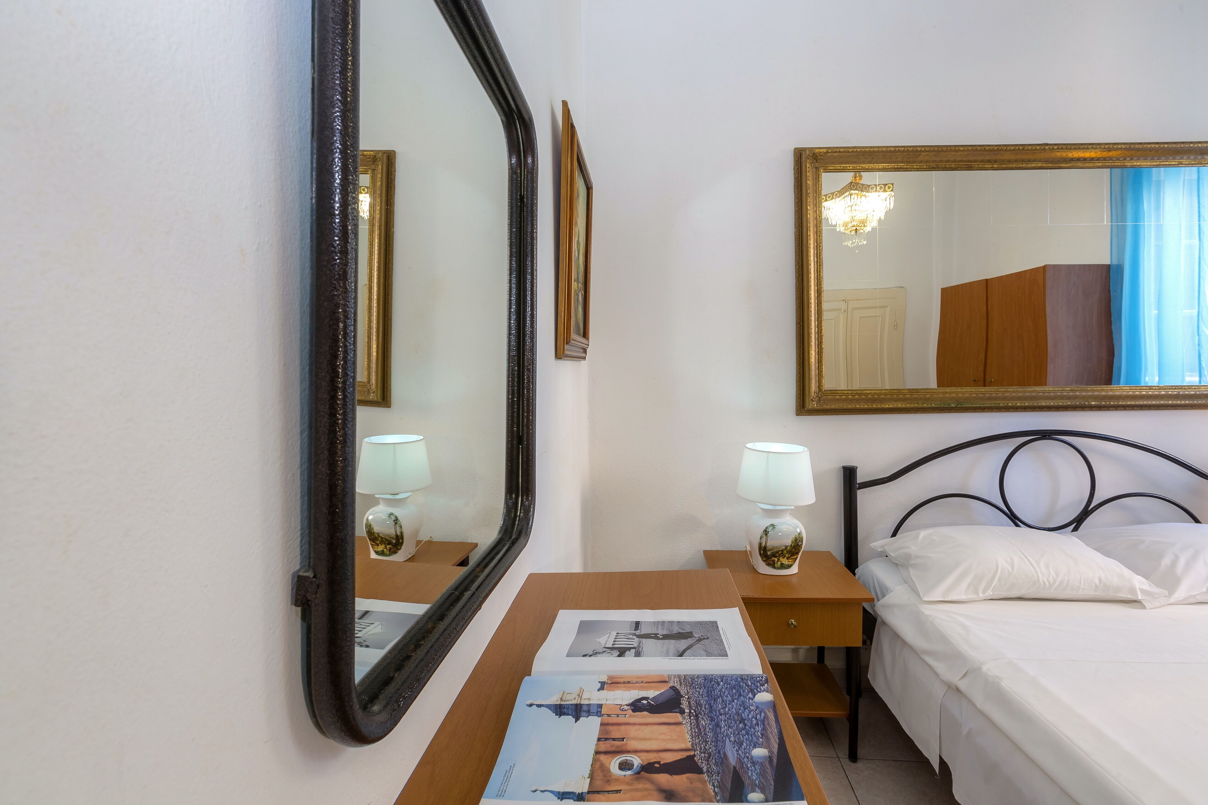 Habitación de hotel con un espejo grande, una cama con ropa blanca, una mesita de noche de madera con una lámpara, y un cuadro enmarcado en la pared.