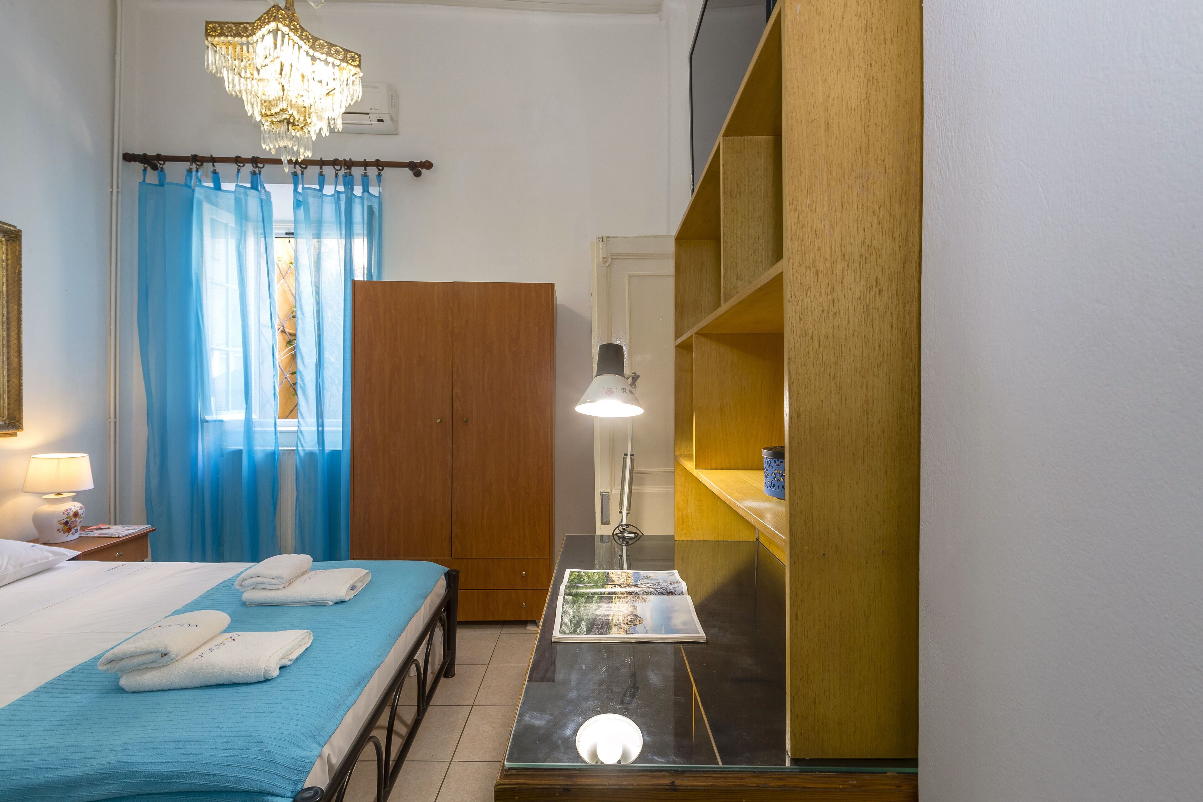 A hotel room with a bed, a desk, a lamp, a blue curtain, and a wooden wardrobe.