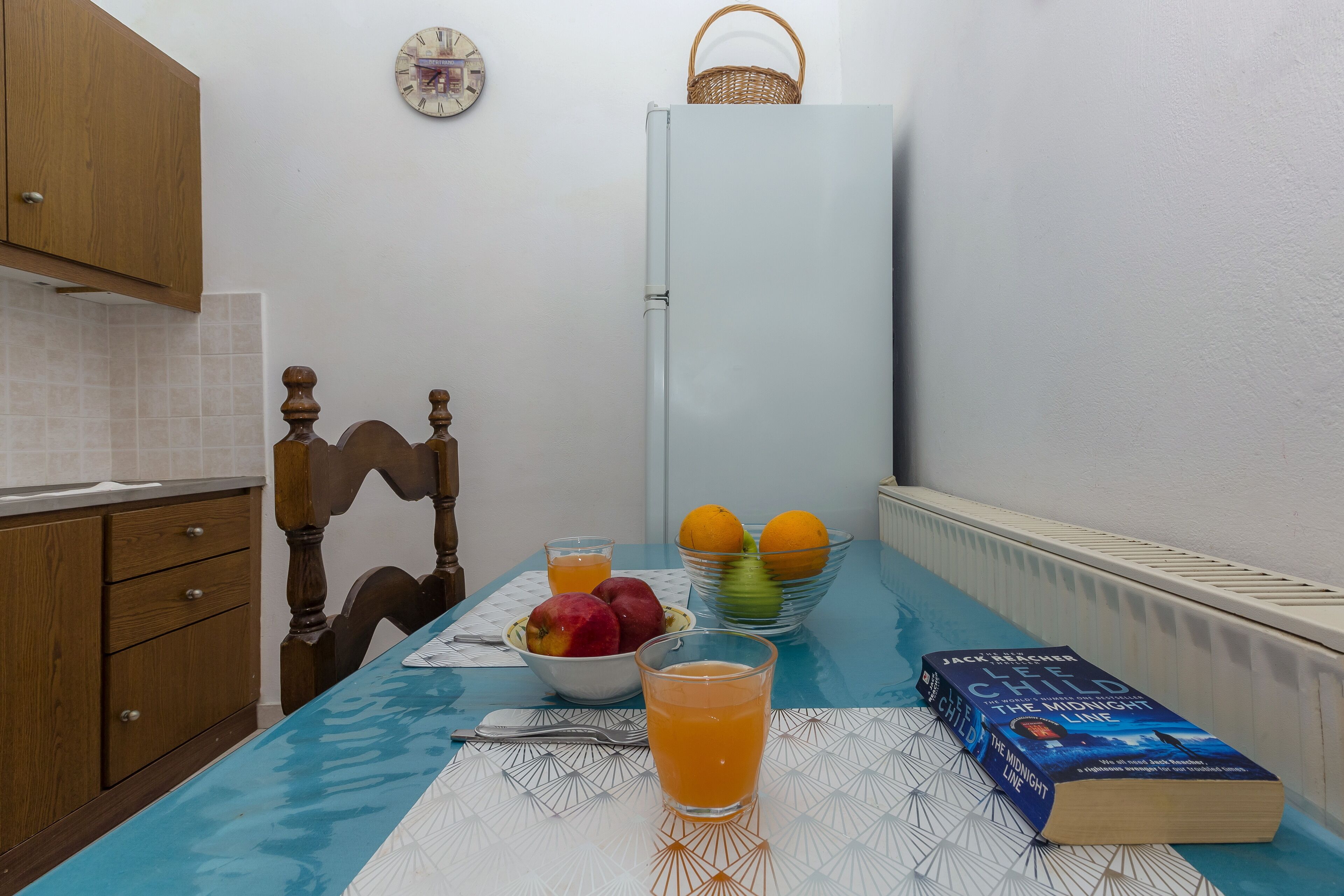 A kitchen with a dining table set for two, a refrigerator, a clock, and a book titled "The Midnight Line".