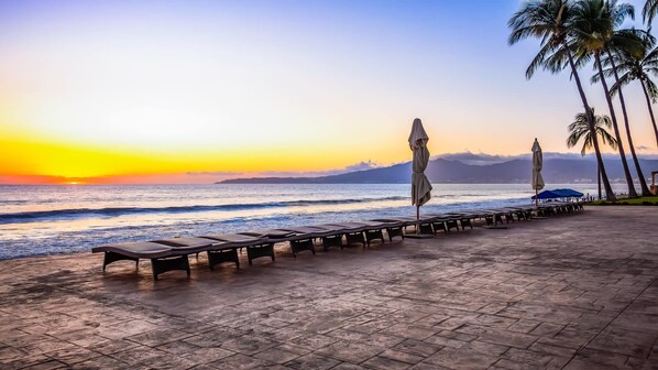 A beachside lounge with sun loungers and umbrellas, overlooking the ocean and a sunset.