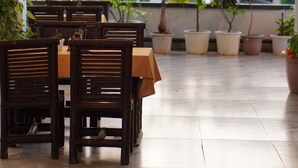 A restaurant with wooden tables and chairs, potted plants, and a view of greenery and buildings in the background. - Club Nirvana Boutique Resort Pure-Veg (Sonapur)