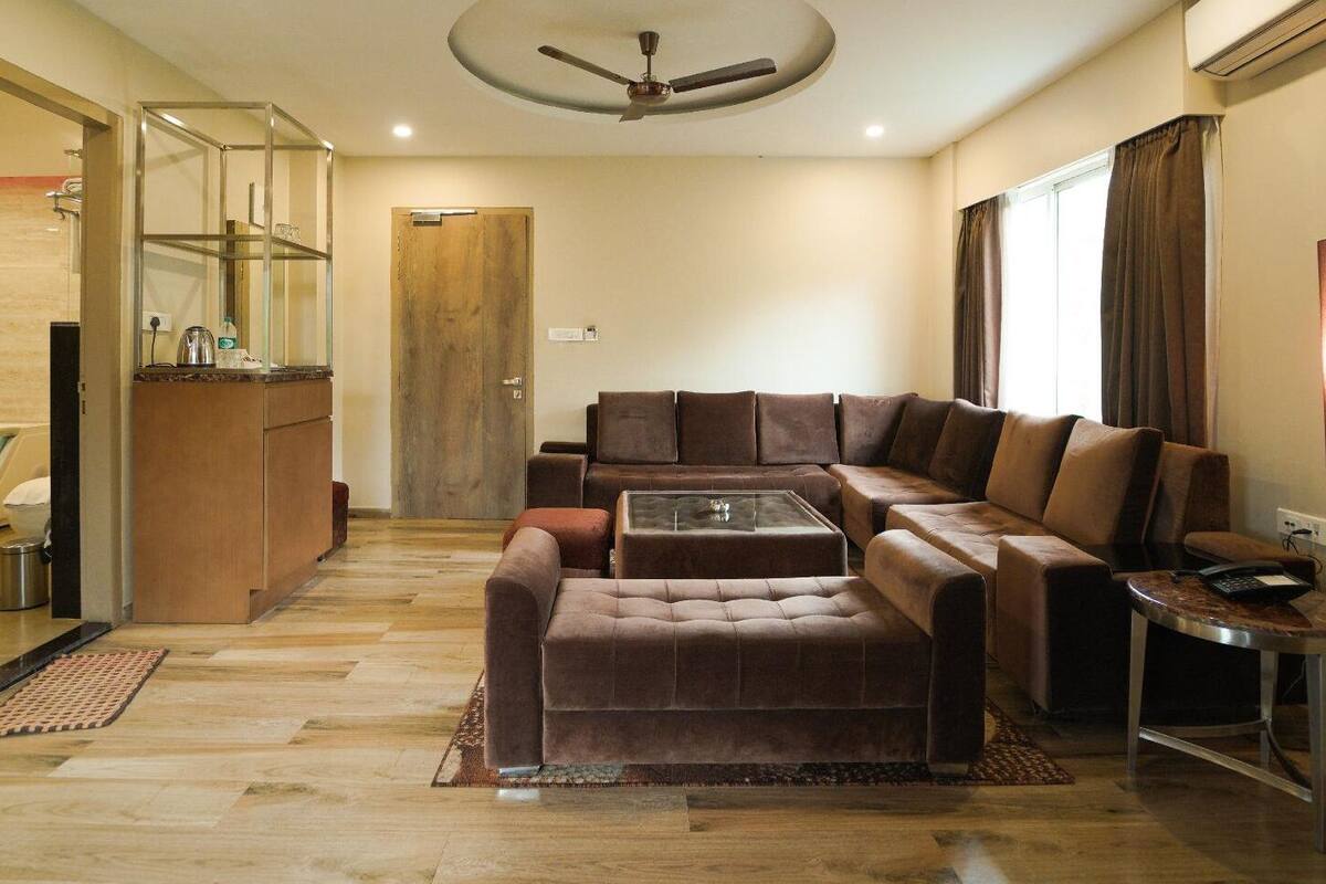 A modern living room with a sectional sofa, a coffee table, and a small side table. There is a wooden door, a ceiling fan, and a window with curtains.