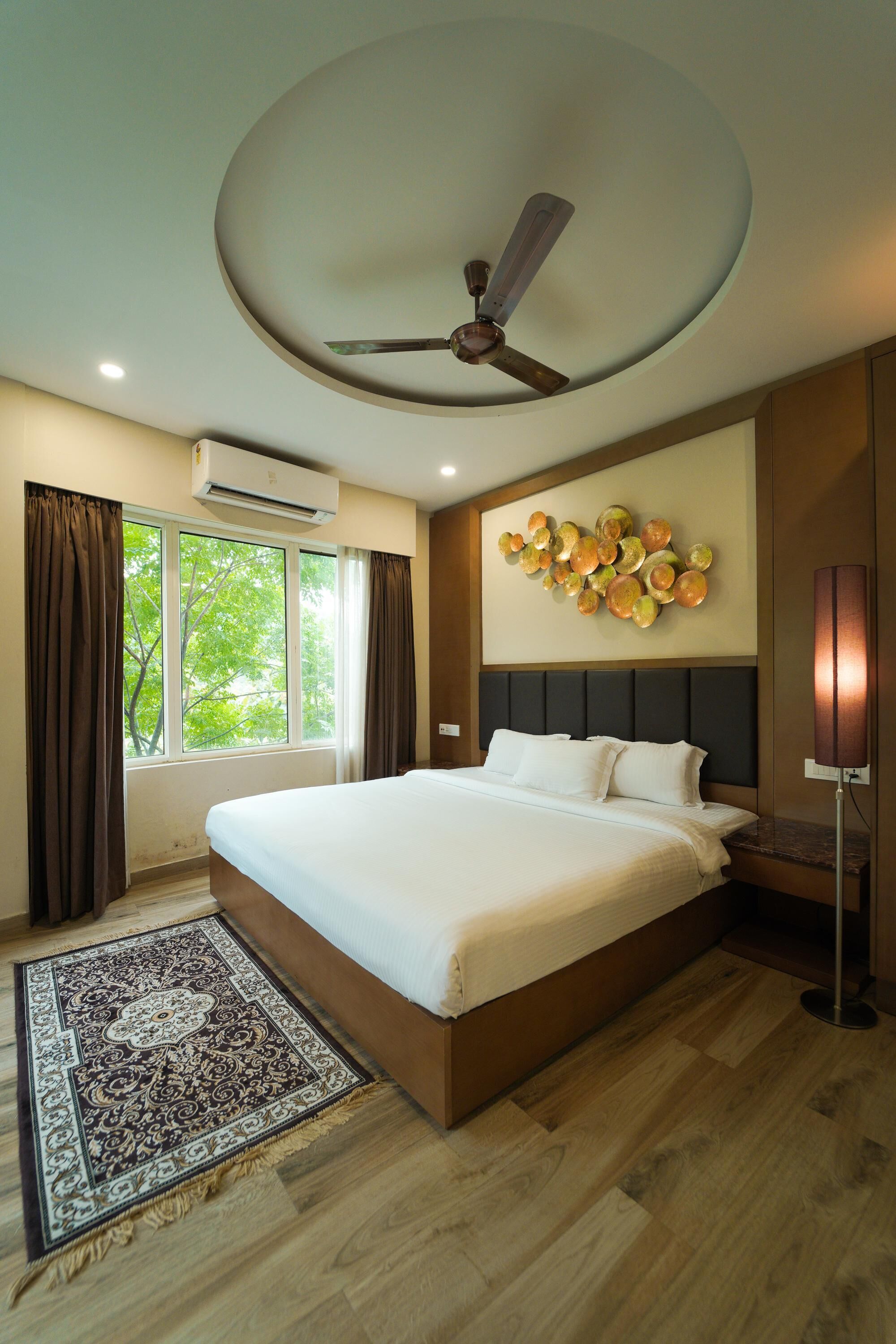 A modern hotel room with a large bed, a ceiling fan, an air conditioning unit, a decorative wall piece, and a view of trees through the window.