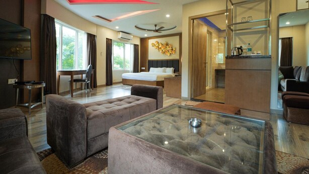 A modern hotel room with a glass coffee table, plush seating, a bed, a dining area, and a kitchenette. - Club Nirvana Boutique Resort Pure-Veg (Sonapur)