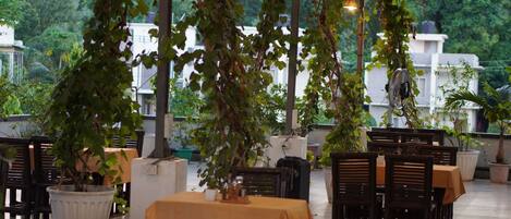 Un área de comedor al aire libre con mesas y sillas, rodeada de plantas en macetas y árboles.