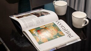 Una revista abierta sobre una mesa negra brillante, una taza blanca y un jarrón azul con flores.