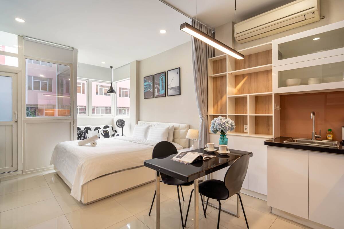 A modern hotel room with a bed, a dining table with chairs, a kitchen area, and a view of the outside.