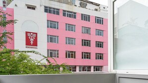 Un balcón con dos taburetes negros, vista de un edificio rosa y blanco con un cartel de hospital, y una ventana con vidrio esmerilado.