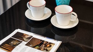 Una mesa con dos tazas blancas de borde rojo, un jarrón azul con flores azul claro y una revista sobre la mesa.