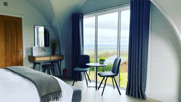 A bedroom with a bed, a desk, a chair, and a view of the countryside through large windows.
