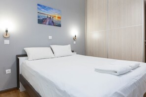 A neatly made bed with white linens and pillows, a painting of a sunset on the wall, and a wooden wardrobe. - Modern Cozy 1 Bd Piraeus Perfection Apt (Piraeus)