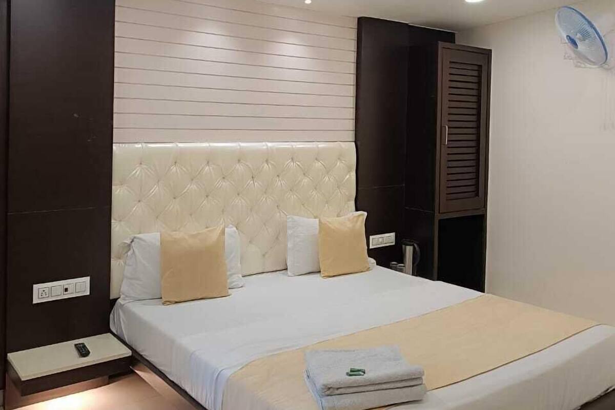 Une chambre moderne avec un grand lit, une tête de lit dotée d’un panneau de couleur claire, une table de chevet avec une télécommande et un ventilateur fixé au mur.