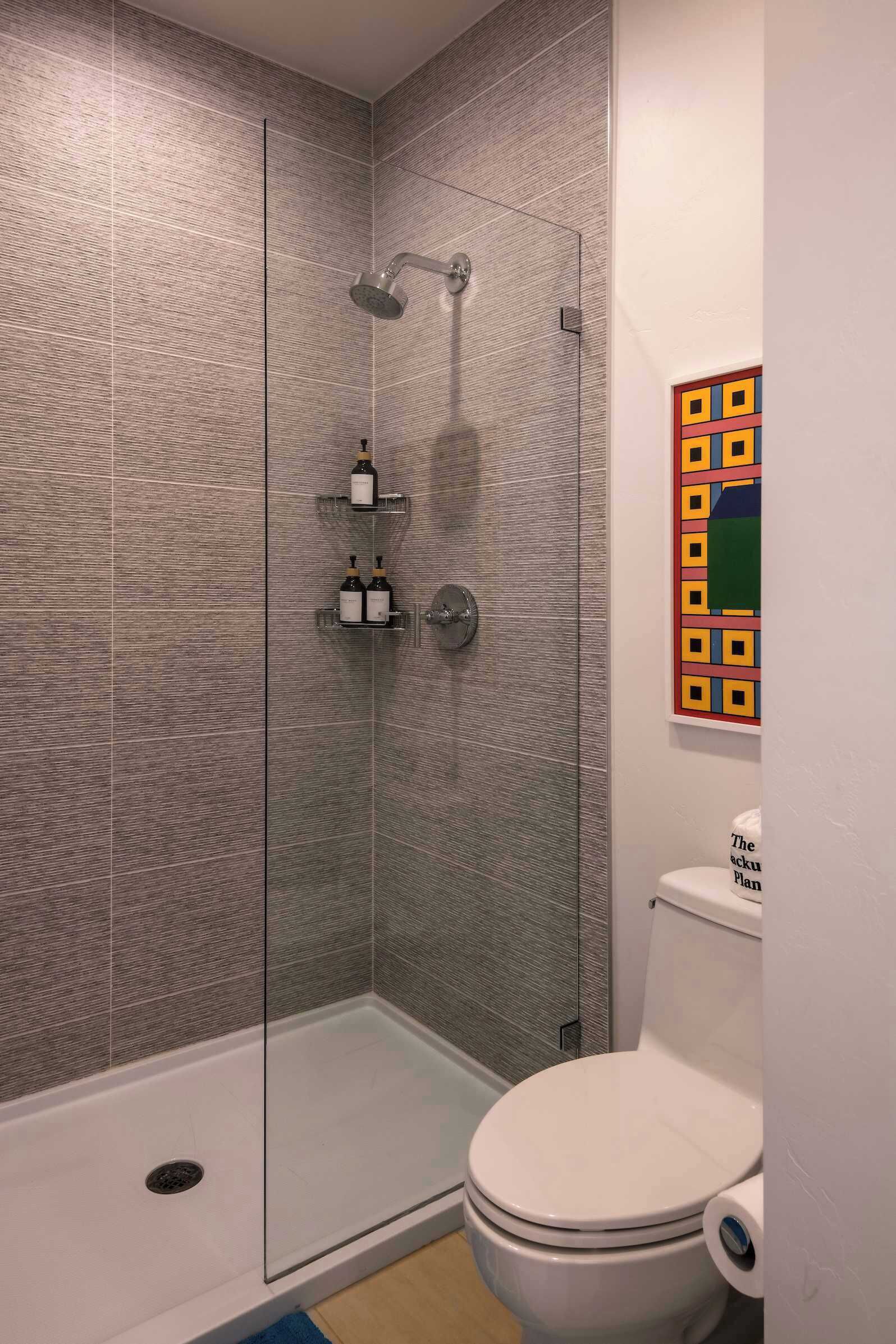 Een moderne badkamer met een douchecabine van glas, een wit toilet en een kleurrijk kunstwerk aan de muur.