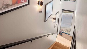 A staircase with a framed poster, wall-mounted light fixtures, and a view of a beach through a window. - Luxury Townhome with Rooftop Spa and Firepit (Steamboat Springs)