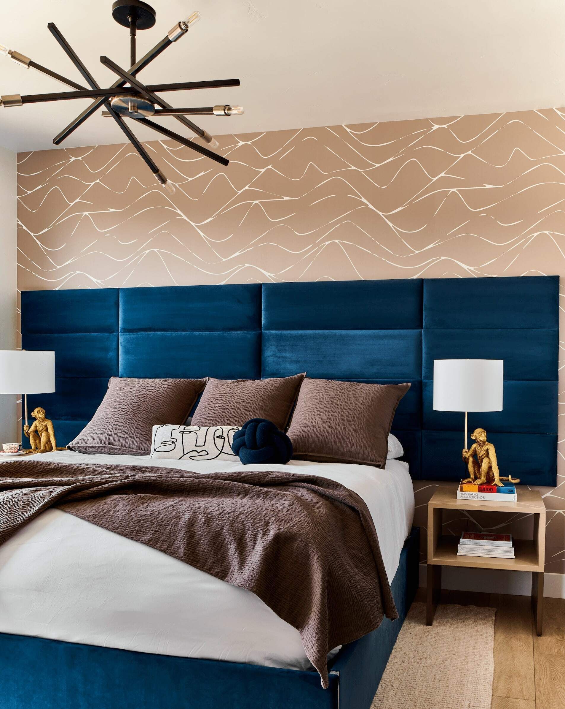 A modern bedroom with a blue upholstered headboard, a patterned wall, and a nightstand with a lamp and books.