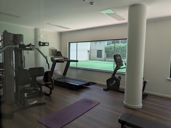 A gym with cardio machines, weight equipment, and a large window offering a view of a tennis court. - Lovely one bedroom city apartment with pool, free parking, gym, walk to the city (Perth)