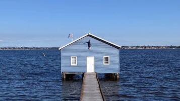 A blue wooden structure with a white door, situated on stilts in the water, connected to land by a wooden dock.