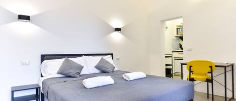 A neatly made bed with a grey bedspread and two pillows, a small bedside table, a yellow chair, and a wall-mounted light fixture.