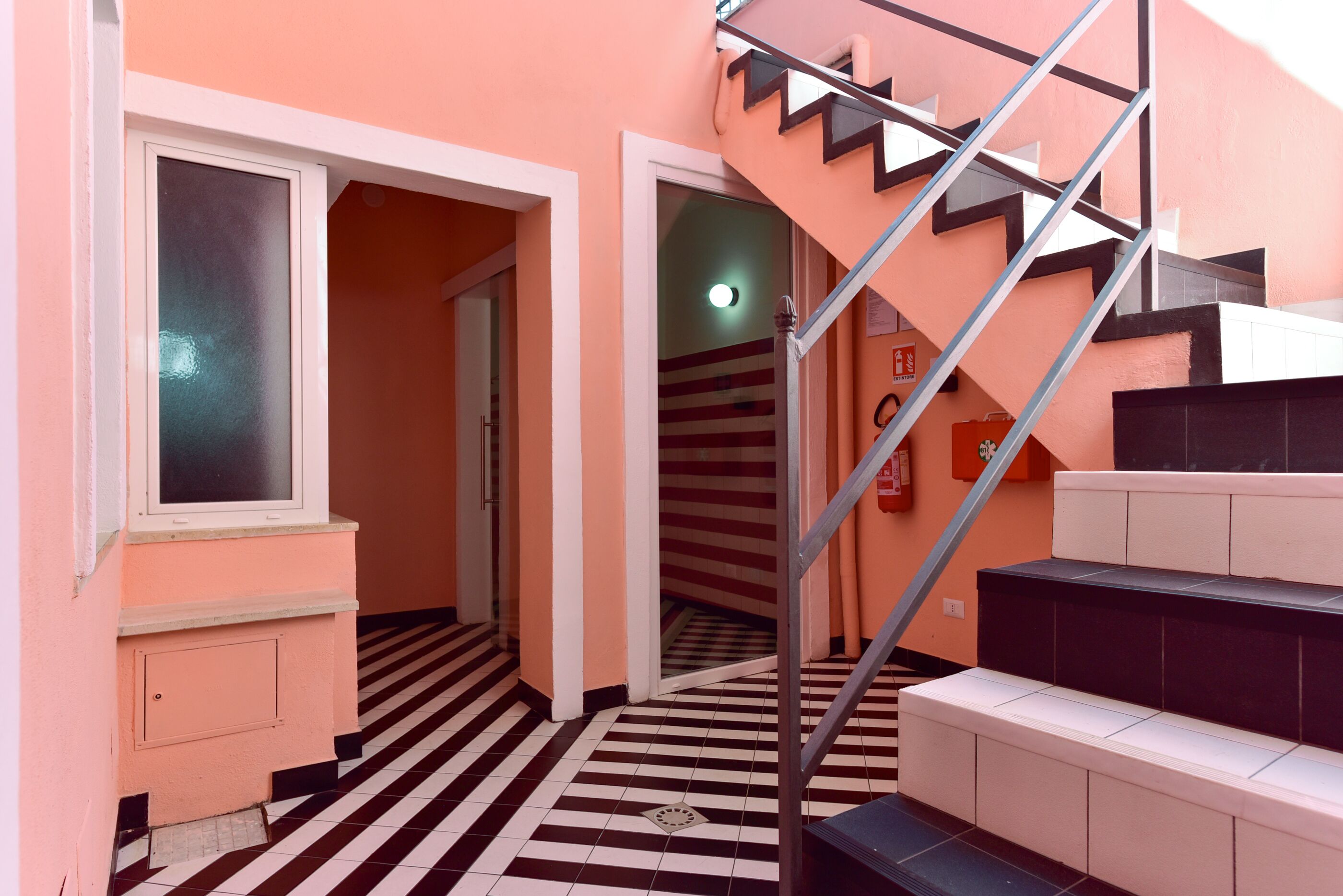 Un pasillo con suelo a cuadros blancos y negros, una escalera con escalones blancos y negros, y una puerta que da a otra habitación.