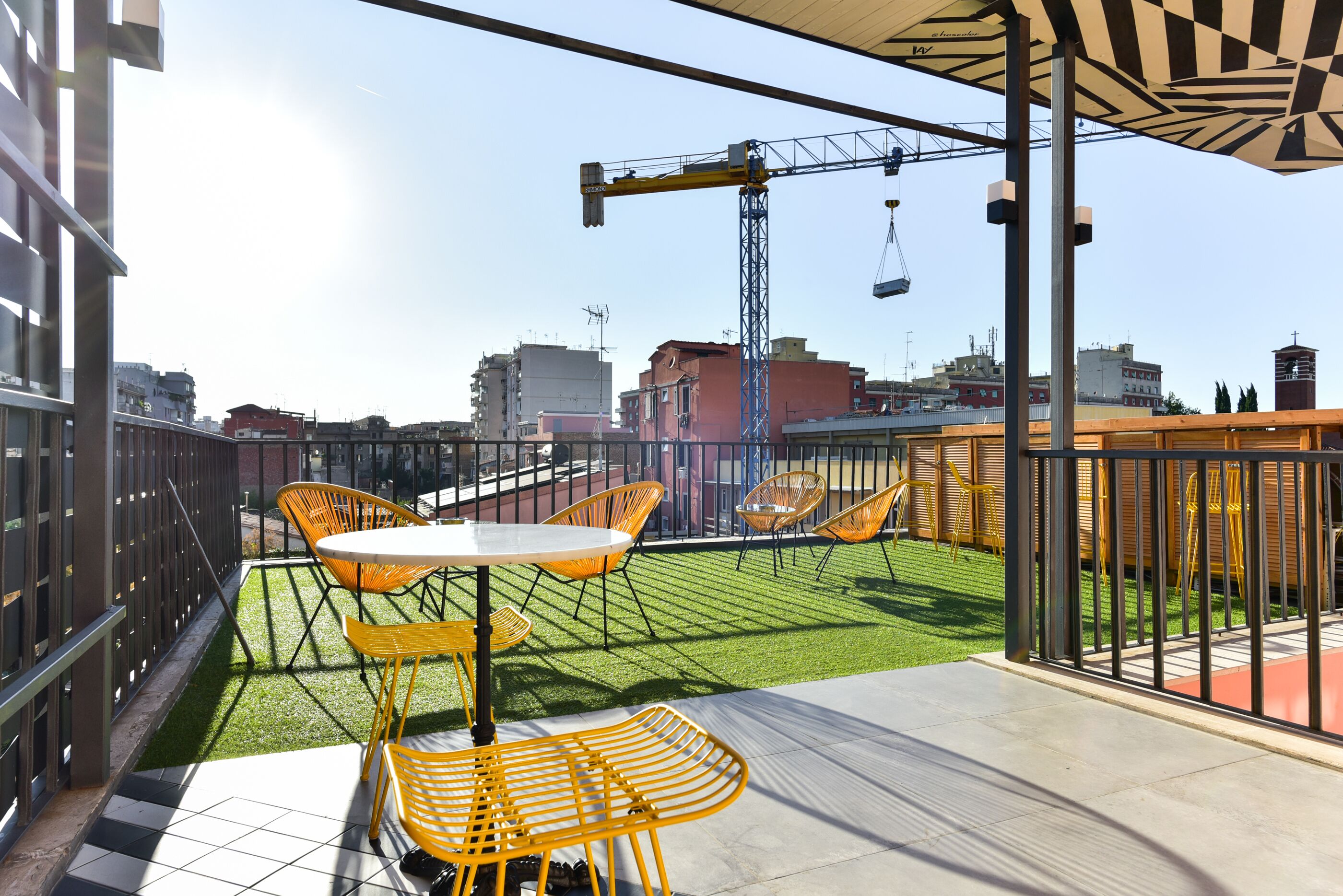 Una terraza en la azotea con sillas amarillas y una mesa, una grúa al fondo y vistas de la ciudad.