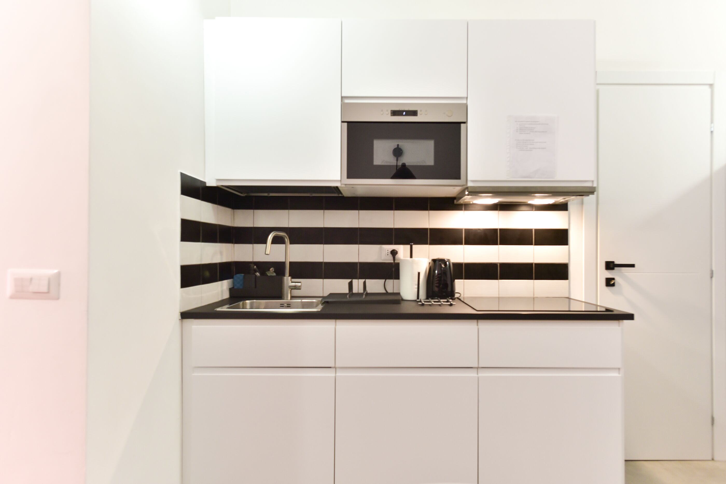 A compact kitchen with white cabinets, a black and white tiled backsplash, a stainless steel sink, and a built-in oven.