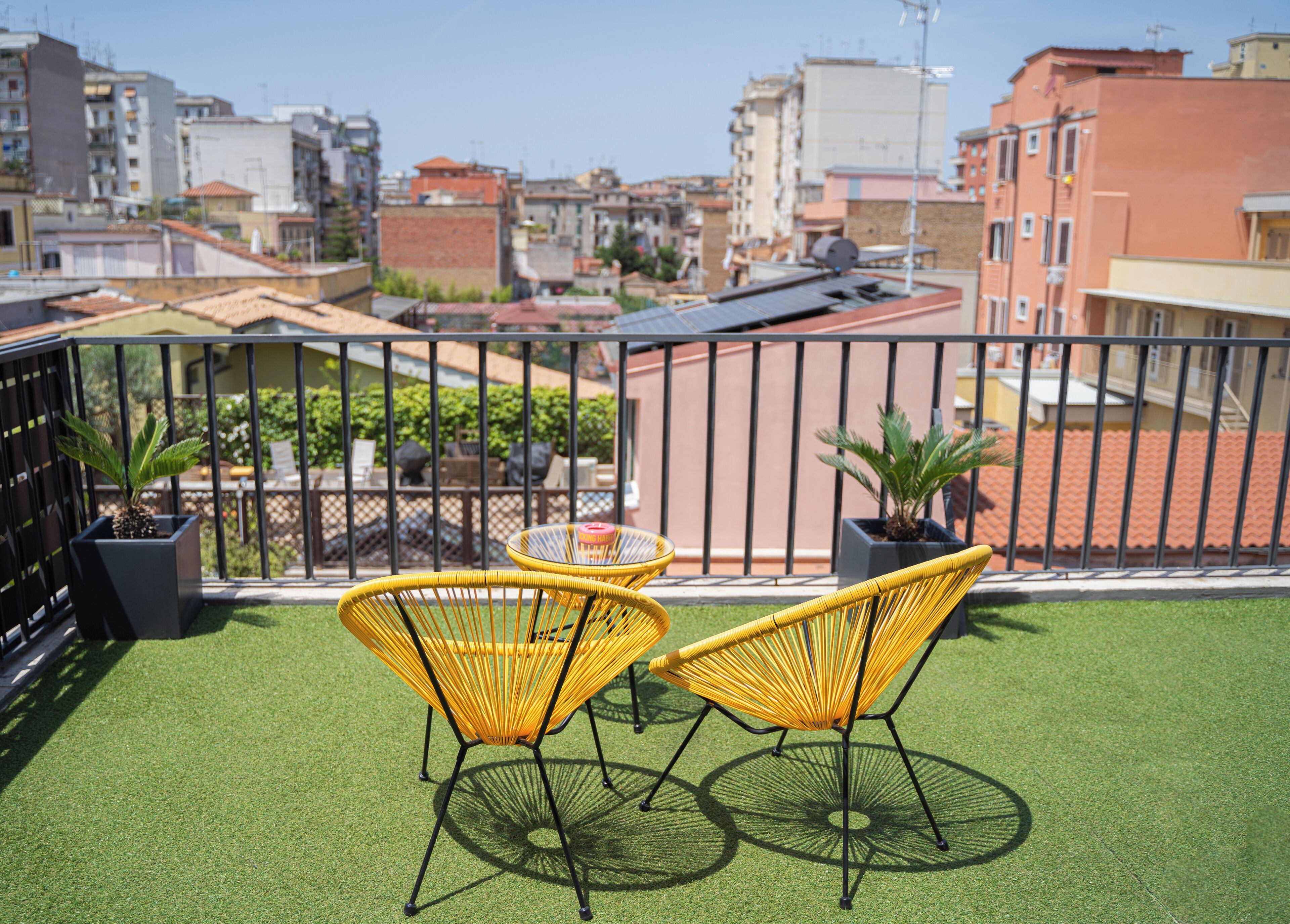 Een balkon met kunstgras, twee gele rieten stoelen, een klein rond tafeltje en potplanten.